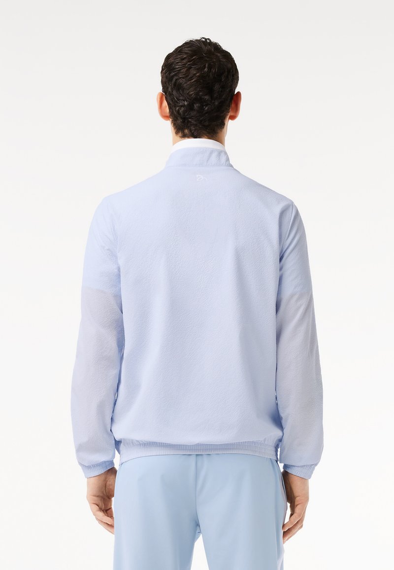 Homme portant une veste texturée bleu clair et un pantalon assorti, se tenant de dos sur un fond blanc uni.