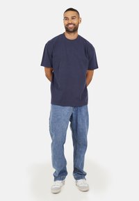 T-shirt en coton bleu marine à col rond, associée à un jean en denim bleu clair. Le modèle se tient les mains derrière le dos, portant des baskets blanches.