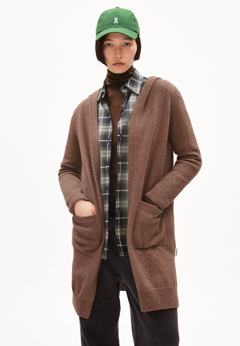 Brauner Strickcardigan mit Kapuze, langen Ärmeln und vorderen Taschen. Über einem karierten Hemd getragen, kombiniert mit einer grünen Mütze.