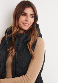 Black quilted puffer vest with a high neck and zip closure, layered over a beige ribbed long-sleeve top. Smooth texture and casual design.