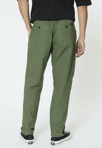 Pantalones cargo verdes con cintura elástica, dos bolsillos traseros con botones y un corte relajado. Combinados con zapatillas negras.