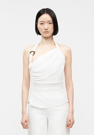 Nainen, jolla on suorat mustat hiukset, pukeutuneena valkoiseen epäsymmetriseen halterneck-toppiin, jossa on koristeellinen vyönsolki yhdellä olkaimella, yhdistettynä valkoisiin housuihin.