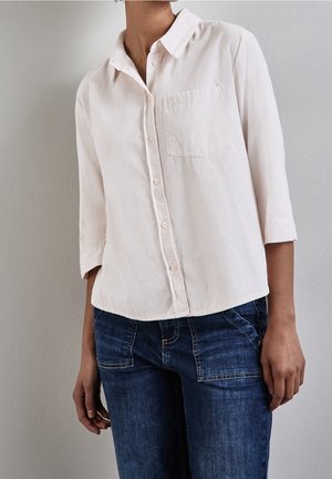 Femme portant une chemise beige clair à boutons avec une poche poitrine et un jean en denim bleu foncé, debout contre un mur gris clair uni.