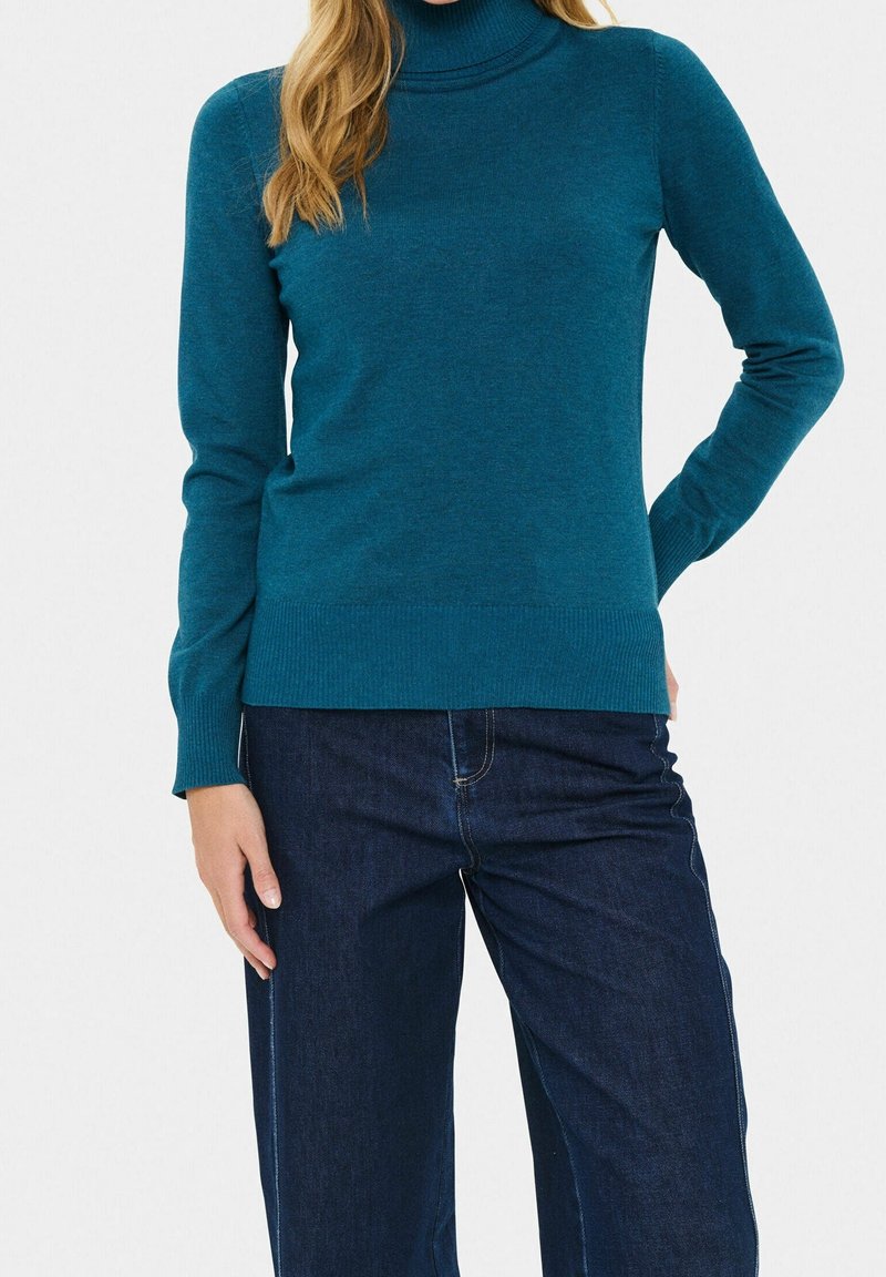 Femme portant un pull à col roulé bleu sarcelle et un jean bleu foncé, debout avec une main sur la hanche sur un fond uni.