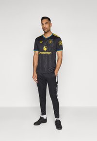 Fekete sportmez rövid ujjakkal, sárga és kék kiegészítőkkel, valamint logóval. Fekete, slim-fit nadrág fehér csíkokkal és fekete sportcipőkkel.