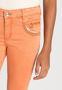 Jeans orange, coupe slim, avec une fermeture éclair sur la poche avant accentuée par du matériel doré et des coutures colorées. Texture douce et lisse.
