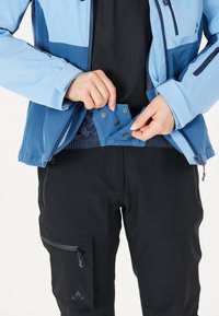 Blå utomhusjacka med kontrasterande mörkblå paneler, dragkedjestängningar och tryckknappar. Kombinerad med svarta byxor som har fickor med dragkedja.