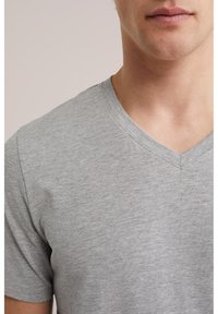 Mladá osoba oblečená v jednoduchom sivom tričku s výstrihom do V a krátkymi rukávmi, zobrazená od stredu hrudníka po bradu na neutrálom pozadí.