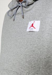 Felpa grigia con tessuto strutturato, caratterizzata da una grande toppa bianca che mostra un logo Jumpman rosso e la scritta "Flight". Dettaglio del cappuccio con lacci.