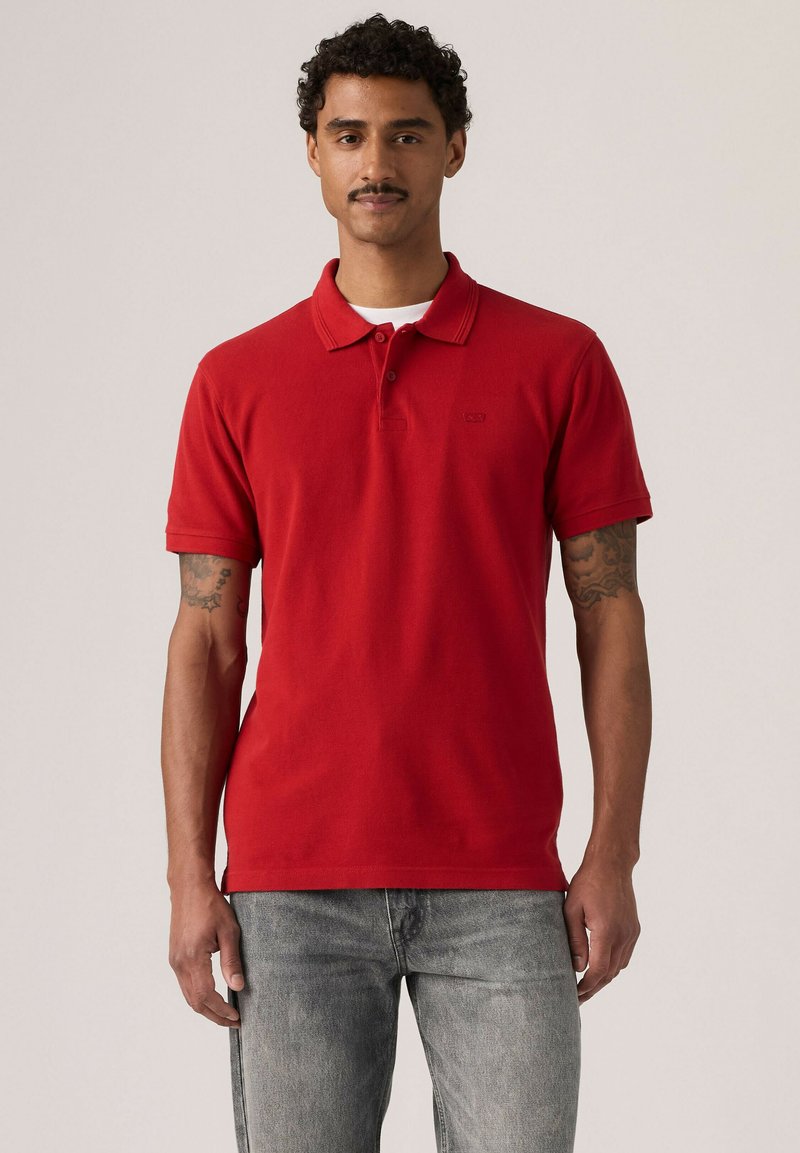 Homme aux cheveux bouclés et moustache porte un polo rouge à manches courtes et un jean gris clair, debout devant un fond clair uni.