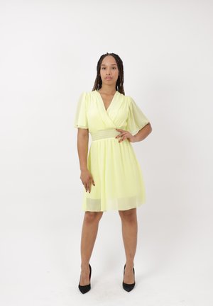 Femme debout dans une robe jaune pâle, longueur genou, à manches courtes bouffantes et talons noirs sur un fond blanc uni.