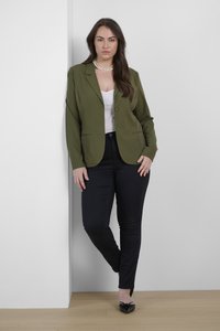 Blazer vert olive avec revers crantés et fermeture à un bouton, associé à un pantalon noir slim fit et des chaussures noires à bout pointu. Tissu lisse.