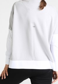 Sudadera gris claro con mangas largas, con un dobladillo blanco y un sutil detalle en la parte trasera. Textura suave, elementos de diseño minimalistas.