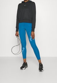 Sudadera negra recortada, leggings ajustados azules con logo blanco de Nike y zapatillas deportivas negras con un acento blanco, sosteniendo una cuerda para saltar.