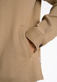 Sudadera beige con un tejido texturizado, que cuenta con un bolsillo lateral y detalles en los puños con botones. La etiqueta de la marca es visible en la parte inferior.