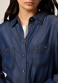 Cămașă din denim închis, cu deschidere cu butoni în față, două buzunare pe piept cu clapete și detalii de cusătură contrastante. Stilul gulerului este clasic.