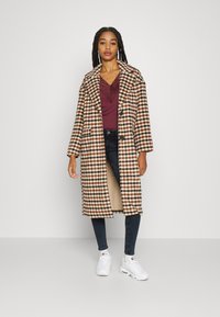 Sobretudo longo castanho e creme com xadrez, com lapelas largas e dois bolsos, usado sobre um top bordô e jeans pretos, combinado com ténis brancos.