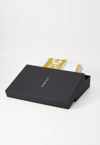 Boîte noire avec le logo "VERSACE", montrant des serviettes blanches avec un motif ornemental doré qui sortent. La boîte a une forme rectangulaire et élégante.