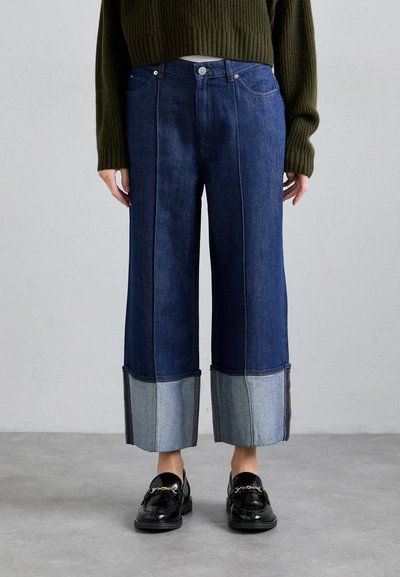 Personne portant un jean large bleu foncé avec de larges revers bleu clair, associé à des mocassins noirs avec un détail de chaîne dorée et un pull vert foncé.