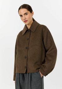 Woman wearing a brown buttoned wool jacket with a wide collar and gray pleated pants, standing with one hand in pocket.