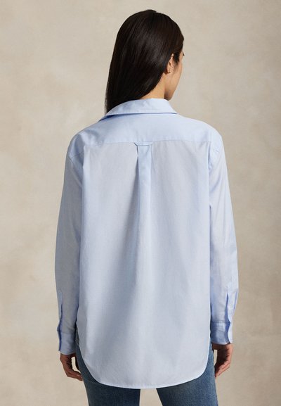 Cămașă albastru deschis cu mâneci lungi, guler largit, pli în spate și tiv curbat. Are manșete cu nasturi și textură netedă.