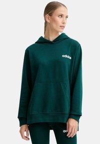 Donna che indossa un felpa Adidas verde scuro e pantaloni abbinati, con lo sguardo rivolto a destra su uno sfondo bianco semplice.