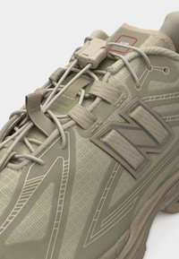 Gros plan d'une chaussure de sport beige avec des motifs complexes, des lacets élastiques, un mécanisme de verrouillage en plastique et un grand logo "N" sur le côté.