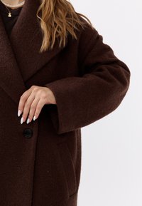 Brauner, übergroßer Wollmantel mit Reverskragen, einem einzelnen schwarzen Knopf und tiefen Taschen. Die strukturierte Stoffqualität und die weiten Ärmel sind sichtbar.