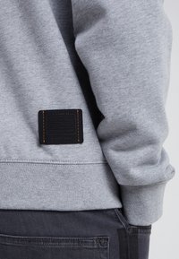 Grå sweatshirt ärm och rygg med svart läder Coach-märke, tillsammans med mörka denimjeans som visar detaljer på bakfickan.