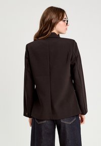 Blazer noir au design structuré, fabriqué en tissu lisse, avec des manches longues et une seule fente à l'arrière. Porte avec un jean sombre.