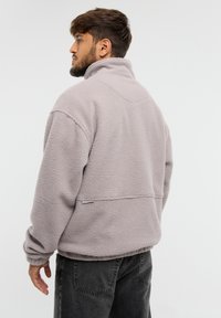 Szary polarowy golf z wysokim kołnierzem, luźny krój, fakturowana powierzchnia i brak widocznych zapięć. Posiada subtelny detal z logo z tyłu.