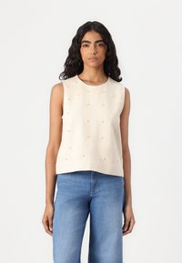 Top senza maniche crema con decorazioni testurizzate che somigliano a perle, con scollatura rotonda e vestibilità dritta, abbinato a jeans azzurri chiari.
