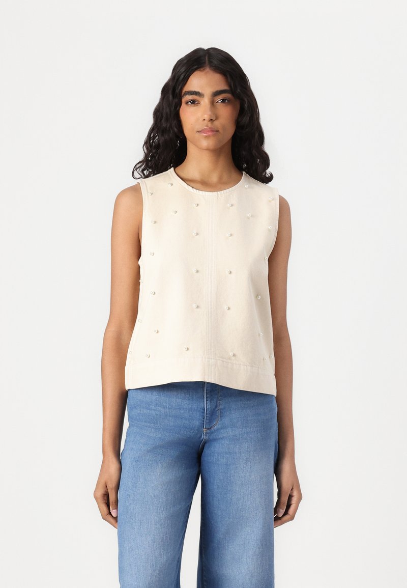 Top senza maniche crema con decorazioni testurizzate che somigliano a perle, con scollatura rotonda e vestibilità dritta, abbinato a jeans azzurri chiari.