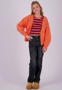 Cardigan arancione oversize lavorato a maglia, top corto a righe rosse e viola, jeans scuri e scarpe platform color cuoio. La modella posa con una mano sul fianco.
