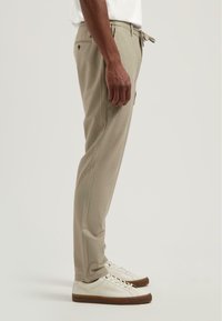 Man in beige op maat gemaakte broek met zij- en achterzakken, witte sneakers met bruine zolen, staand tegen een effen achtergrond.