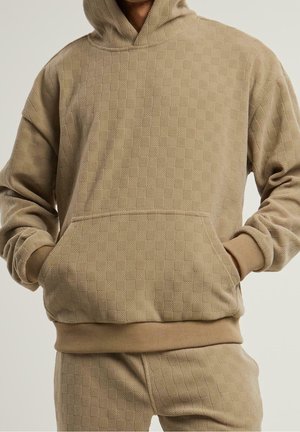 Personne portant un sweat à capuche beige à carreaux texturé et un pantalon assorti, les mains dans la poche avant, sur un fond uni.