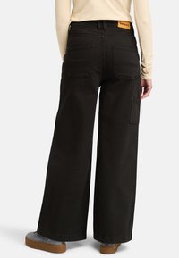 Pantalones cargo negros de pierna ancha con grandes bolsillos traseros y una etiqueta con el logo en la cintura, combinados con calzado gris.
