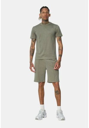 Homme tatoué portant une chemise à manches courtes et un short vert olive, des chaussettes blanches et des baskets grises, debout devant un fond uni.