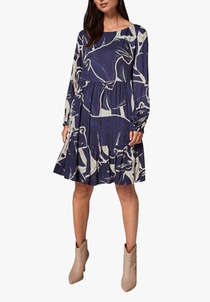 Femme vêtue d'une robe bleu marine à manches longues et longueur genou, ornée d'imprimés abstraits beiges représentant des animaux, et portant des bottines beiges.