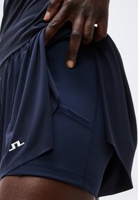 Άτομο σηκώνει μπλε σκούρα φούστα για να αποκαλύψει ταίριασμα σφιχτών σορτς με μικρό λευκό λογότυπο στο κάτω αριστερό μέρος.