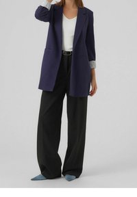 Granatowa marynarka o dopasowanym kroju, z długimi rękawami i podwójnymi kieszeniami z przodu, zestawiona z czarnymi spodniami o szerokich nogawkach i jasnoniebieskimi butami na szpilkach.