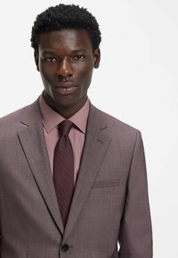 Herrenanzug aus hellviolettem Stoff mit subtiler Struktur, kombiniert mit einem passenden Hemd und einer dunkelvioletten Krawatte. Auffällige Details sind eine Tasche.