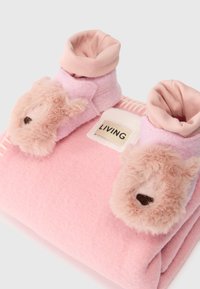 Rozā flīsa čības ar lāča sejām un mīkstā kažokādā. Pagriezti augšēji maliņas gaišākā rozā krāsā un "LIVING" etiķete uz gaiša auduma fona.