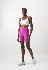 Hvit trenings-bh med svarte detaljer, kombinert med lyserøde tettsittende shorts. Modellen har på seg lyse joggesko med blå og grå detaljer.