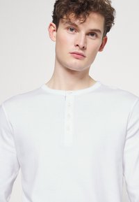 Camicia henley bianca a maniche lunghe con scollatura rotonda, placket a quattro bottoni e texture morbida. Design semplice, senza motivi o accenti.