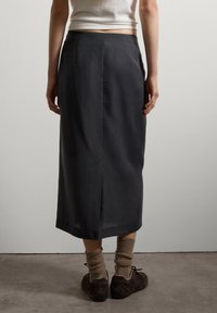 Une personne portant une jupe midi noire avec une fente à l'arrière, des chaussettes brunes, des chaussures marron foncé et un débardeur blanc court, se tenant sur un sol gris.