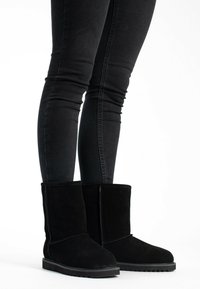 Botas de ante negras hasta el tobillo con punta redondeada, suela de goma plana y detalles de costura simples. Llevadas con jeans negros de corte ajustado.