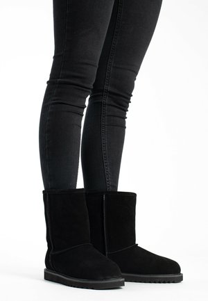 Black suede ankle boots with a round toe, flat rubber sole, and simple stitching details. Worn with black slim-fit jeans.