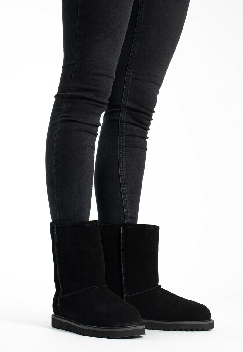 Botas de ante negras hasta el tobillo con punta redondeada, suela de goma plana y detalles de costura simples. Llevadas con jeans negros de corte ajustado.
