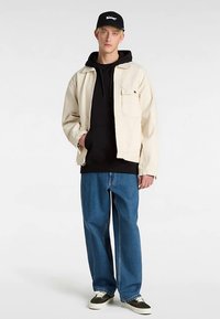 Beige cotton jacket over a black hoodie, paired with blue loose-fit jeans and black-and-white sneakers, with a black cap featuring logo detail.